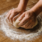 Mâini frământând un aluat pe un blat de lemn presărat cu făină – simbol al aluaturilor de bază pentru pâine, plăcinte și pizza